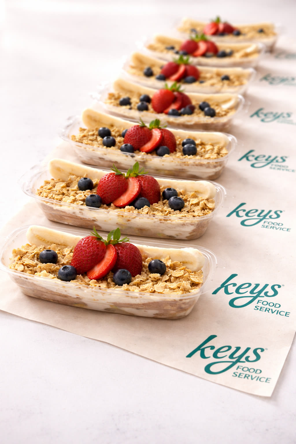 A line of KEYS-branded yogurt and granola parfait cups topped with strawberries, blueberries, and banana on KEYS Food Service parchment