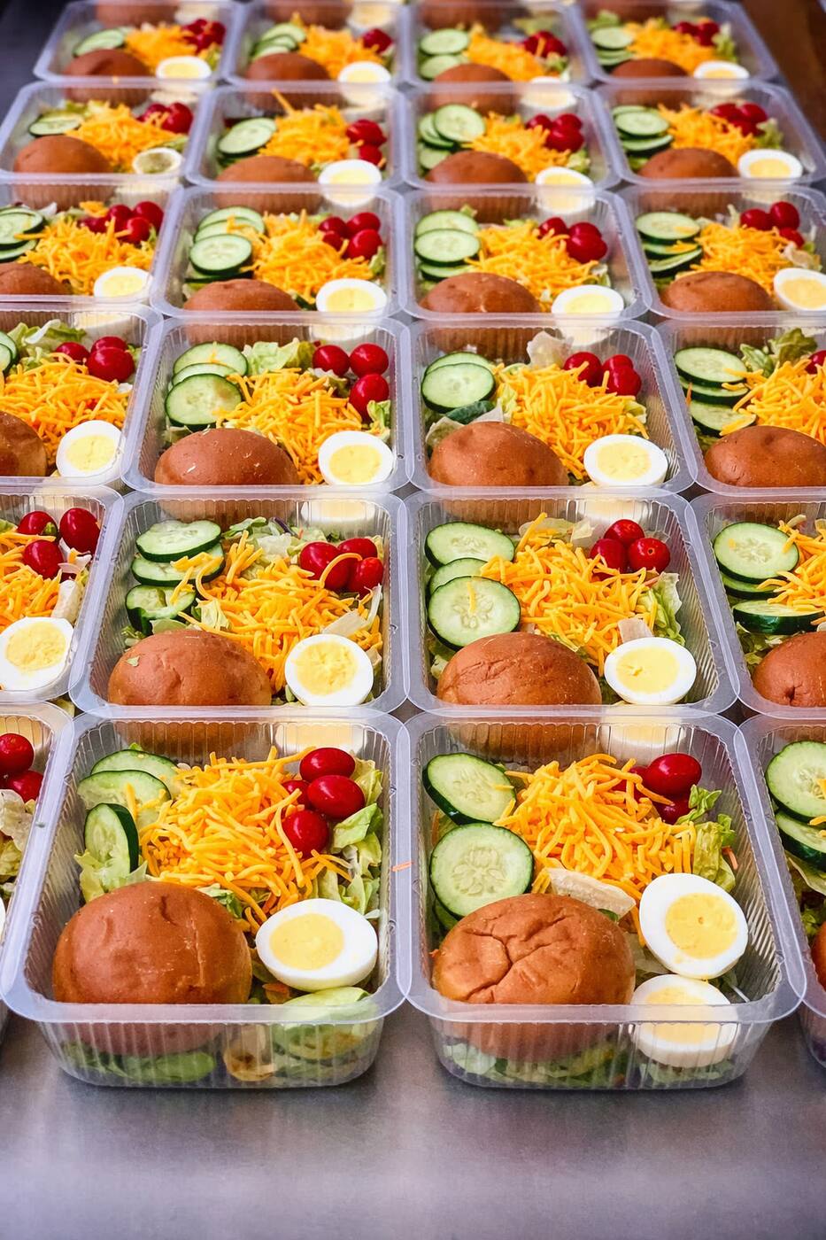 Rows of salad trays with shredded cheddar, cucumber, cherry tomatoes, hard-boiled egg, and a soft brioche bun, packed for daily delivery