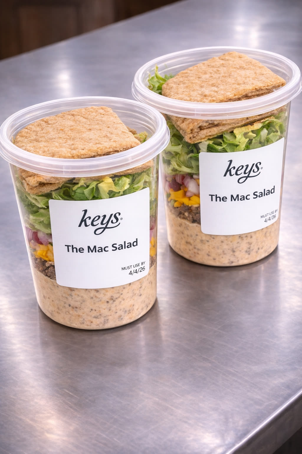 Two KEYS Mac Salad jars on a stainless prep table, each layered with grains, romaine, cheddar and dressing